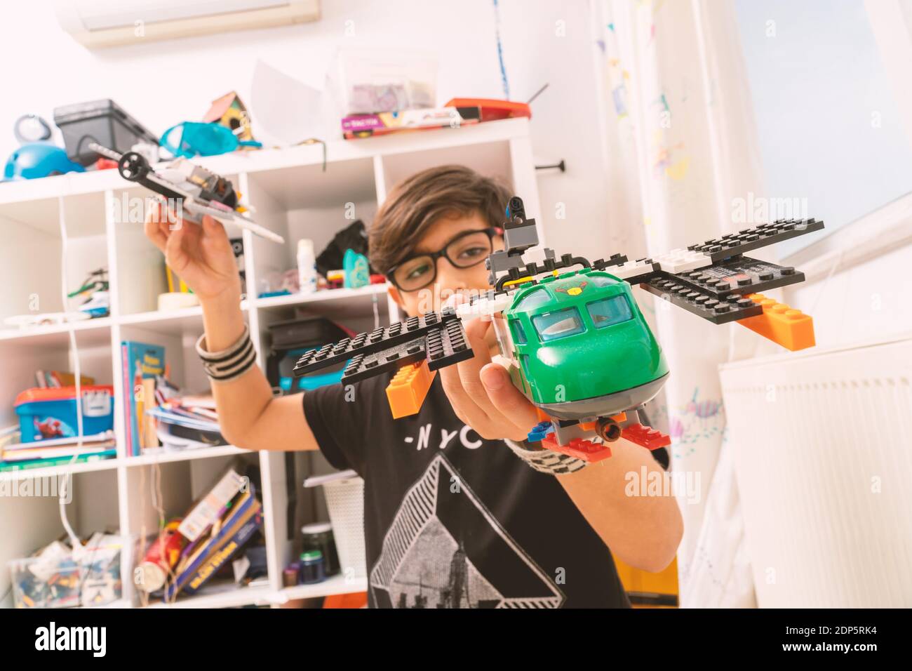 Focus on air craft toy hold by a boy with glasses. The kid is playing alone at home during Covid 9 quarantine. He has a shelf full pf toys. Stock Photo