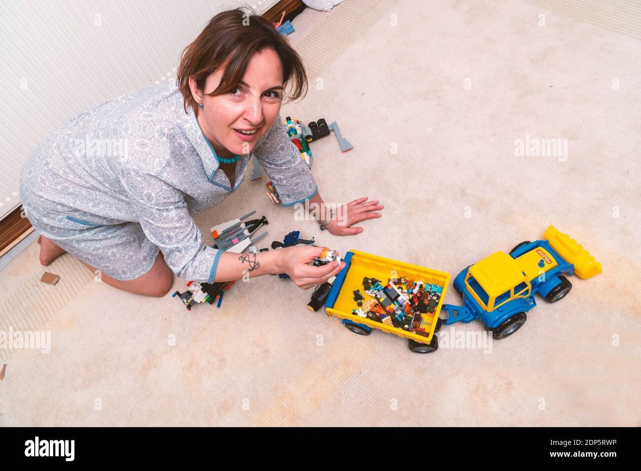 Exhausted and elegant dressed business woman is sitting on the floor and cleaning the toys of her kids. She looks unhappy and frustrated Stock Photo
