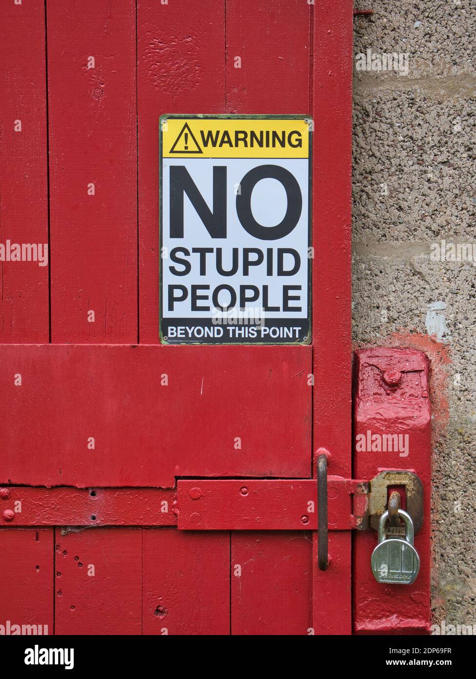 A humorous safety sign saying 'Warning! No stupid people beyond this point' fixed to a red, wooden door locked with a padlock and hasp. Stock Photo