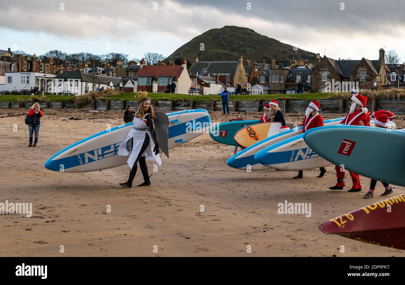 North Berwick, East Lothian, Scotland, United Kingdom, 19th December 2020. Paddle Boarding Santas for charity: a local community initiative by North Berwick News and Views called 'Christmas Cheer' raises over £5,000 funds for families in need, The paddle boarders dressed in Santa costumes carry their paddle boards across the beach with a view of Berwick Law in the background Stock Photo