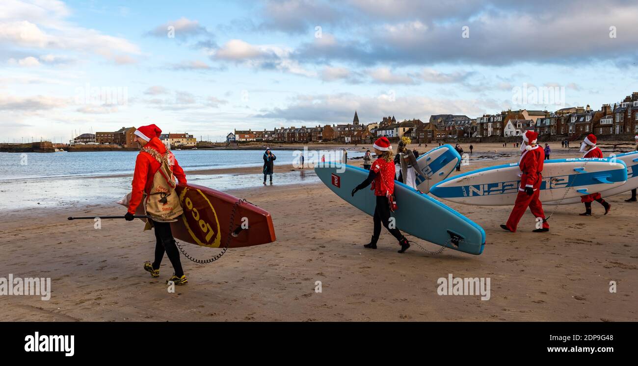 North Berwick, East Lothian, Scotland, United Kingdom, 19th December 2020. Paddle Boarding Santas for charity: a local community initiative by North Berwick News and Views called 'Christmas Cheer' raises over £5,000 funds for families in need, The paddle boarders dressed in Santa costumes carry their paddle boards across the beach Stock Photo