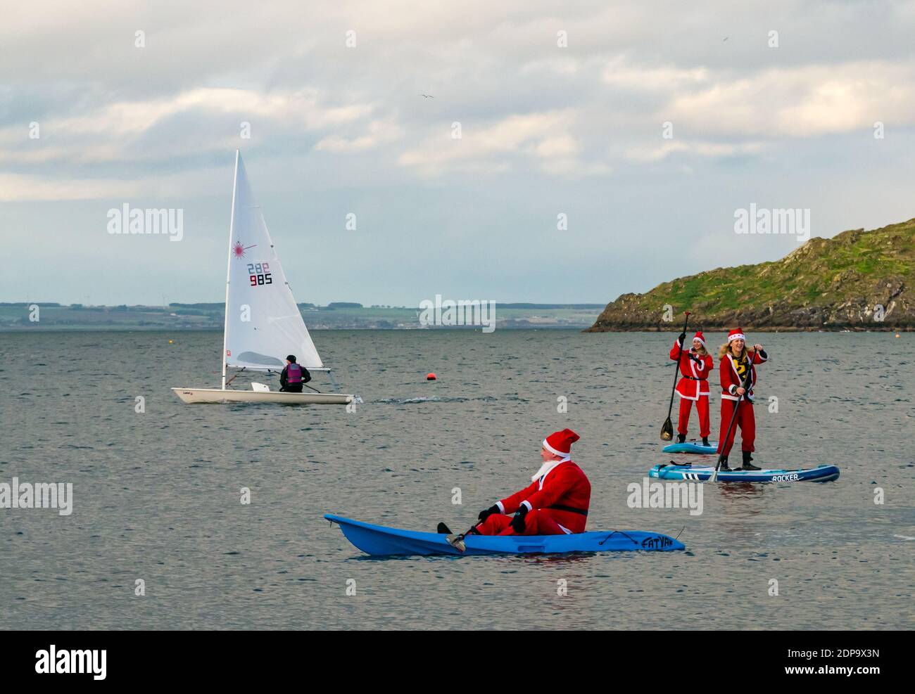 North Berwick, East Lothian, Scotland, United Kingdom, 19th December 2020. Paddle Boarding Santas for charity: a local community initiative by North Berwick News and Views called 'Christmas Cheer' raises over £5,000 funds for families in need. The paddle boarders are dressed in Santa costumes with a sailing boat passing by Stock Photo
