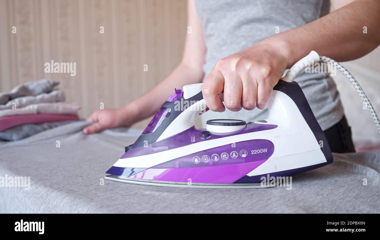 Hardworking woman holding modern purple electric iron irons fast and carefully grey shirt after laundry on special board at home extreme closeup Stock Photo