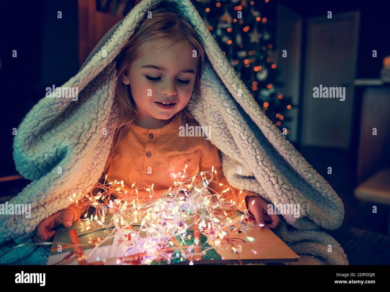 Cute Little Baby Under Plaid with Pleasure Reading Christmas Tale Book and Enjoying Beautiful Glowing Gift. Hygge. Christmas Eve at Home. Stock Photo
