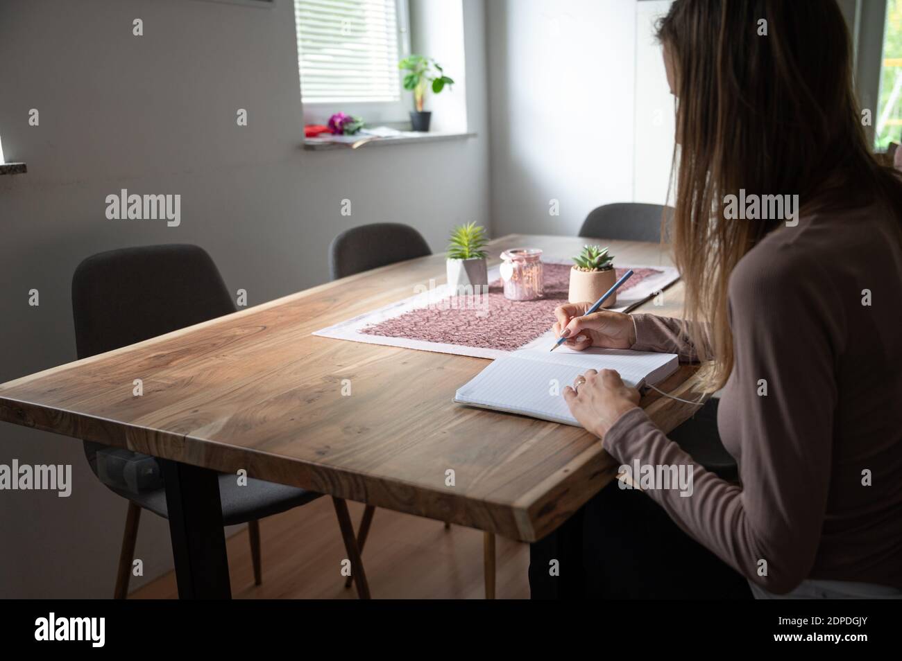 Young woman, a student, sitting at home dining table writing in notepad in a conceptual image of homeschooling or working from home. Stock Photo