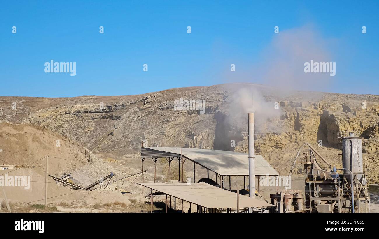 White smoke rises from high chimney on small mining station at large open limestone quarry among hills against blue sky on sunny day Stock Photo
