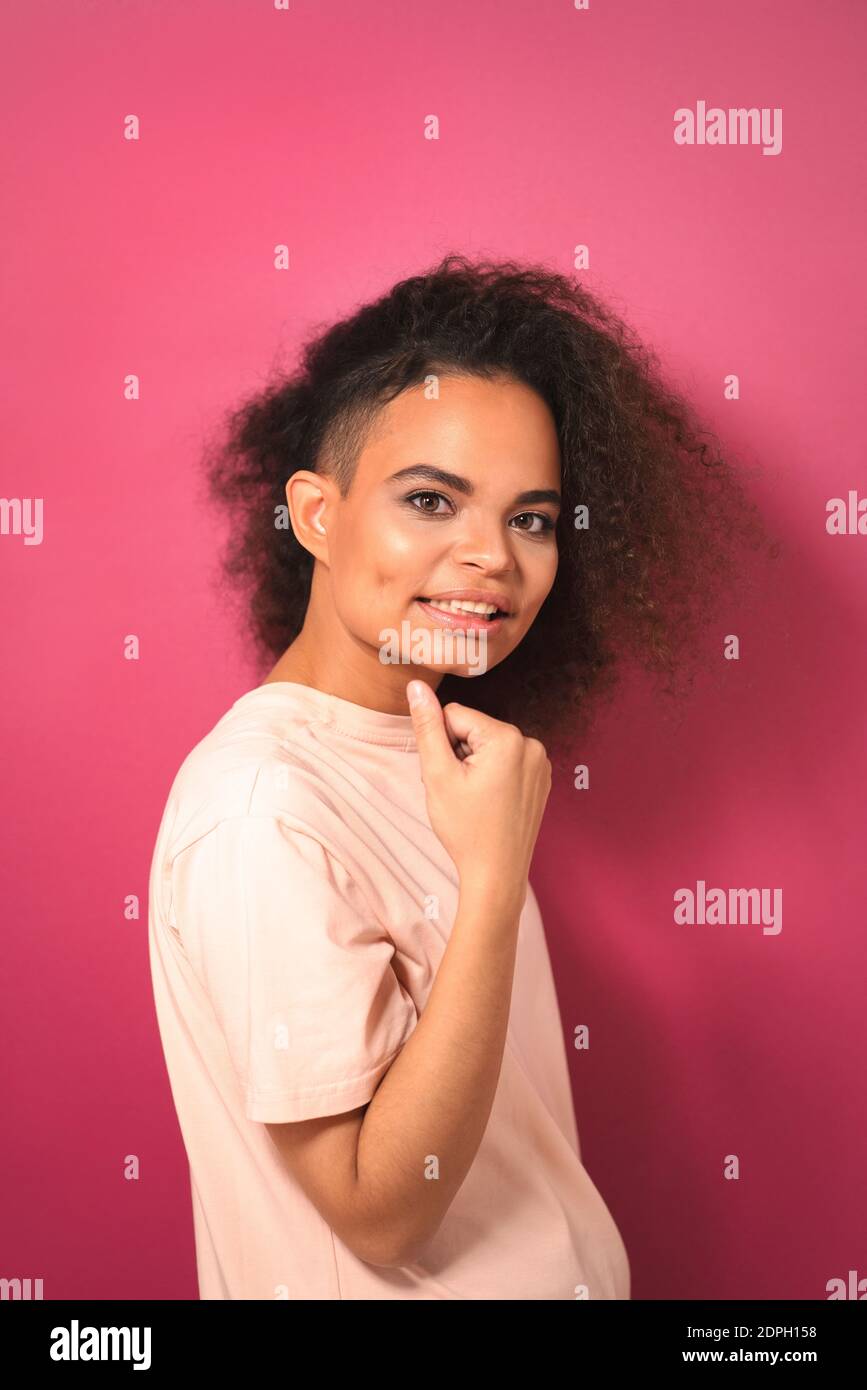 Charming passionate young African American girl with Afro hair looking positively at camera with one arm folded wearing peachy t-shirt isolated on Stock Photo