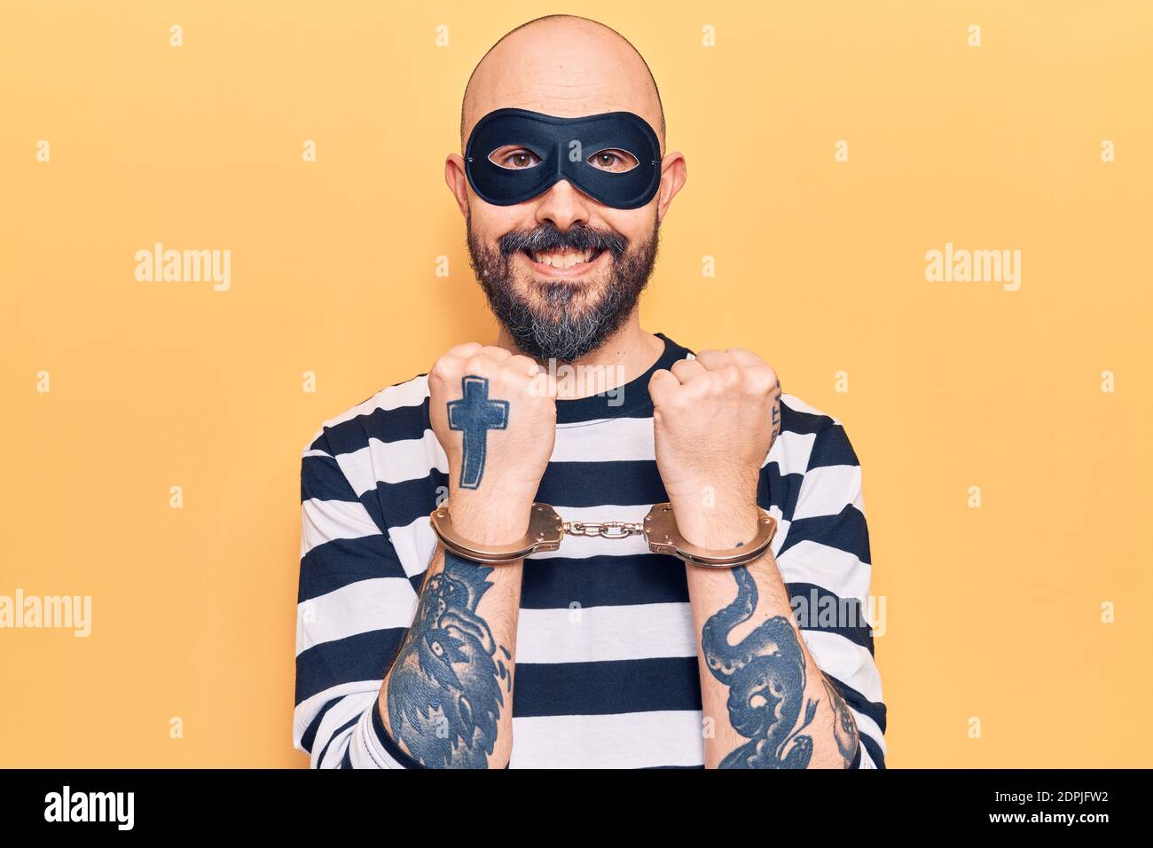Young handsome man wearing burglar mask and handcuffs looking positive and happy standing and smiling with a confident smile showing teeth Stock Photo
