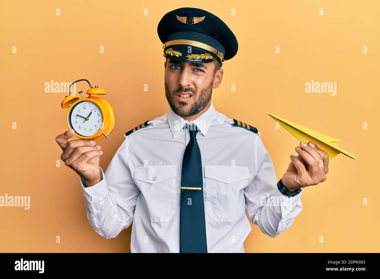 Handsome hispanic pilot man holding paper plane and alarm clock