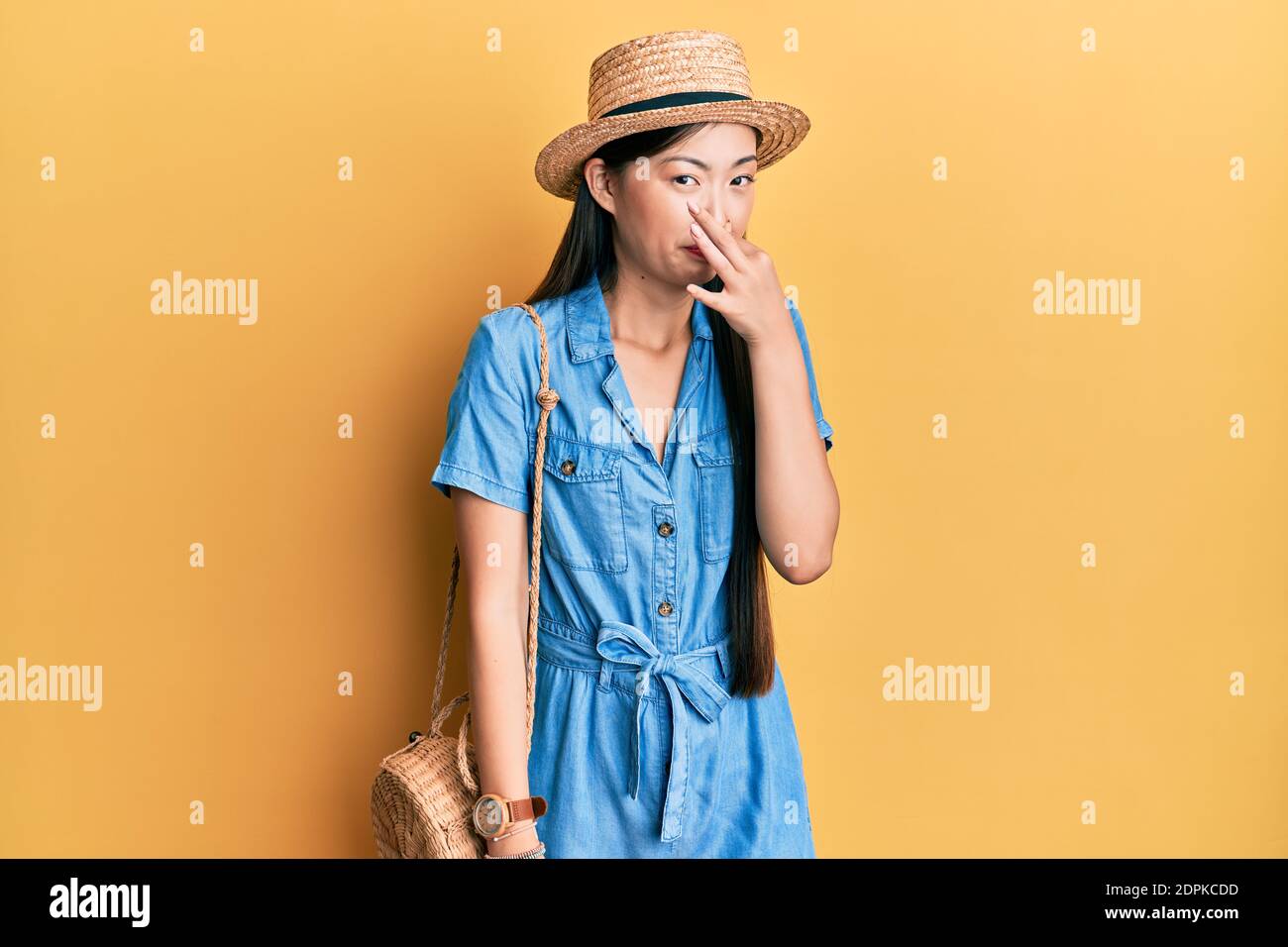 Young chinese woman wearing summer hat smelling something stinky and disgusting, intolerable smell, holding breath with fingers on nose. bad smell Stock Photo