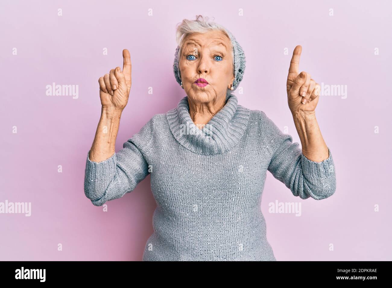 Senior grey-haired woman pointing up with fingers puffing cheeks with funny face. mouth inflated with air, catching air. Stock Photo