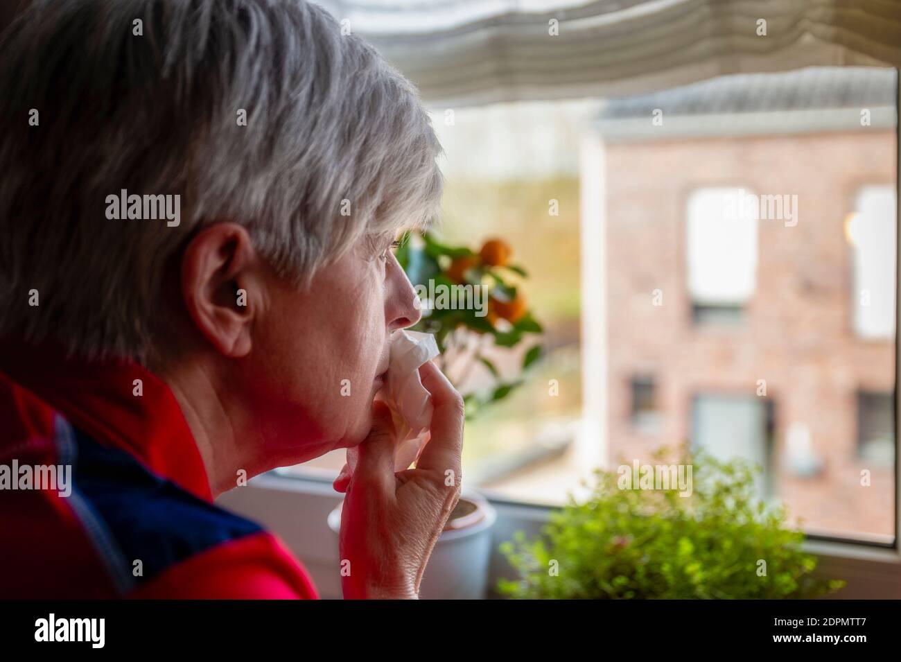 Senior Woman In Quarantine Looks Sadly Out Of The Kitchen Window. Stock Photo