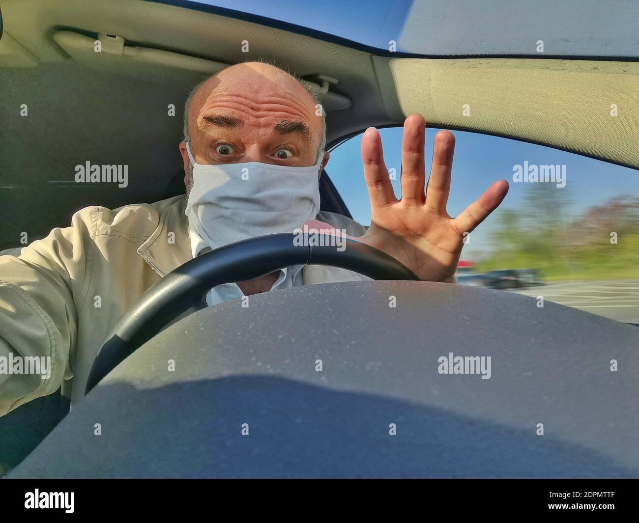 Senior mit Gesichtsmaske und erhobener Hand am Steuer eines PKW. Stock Photo
