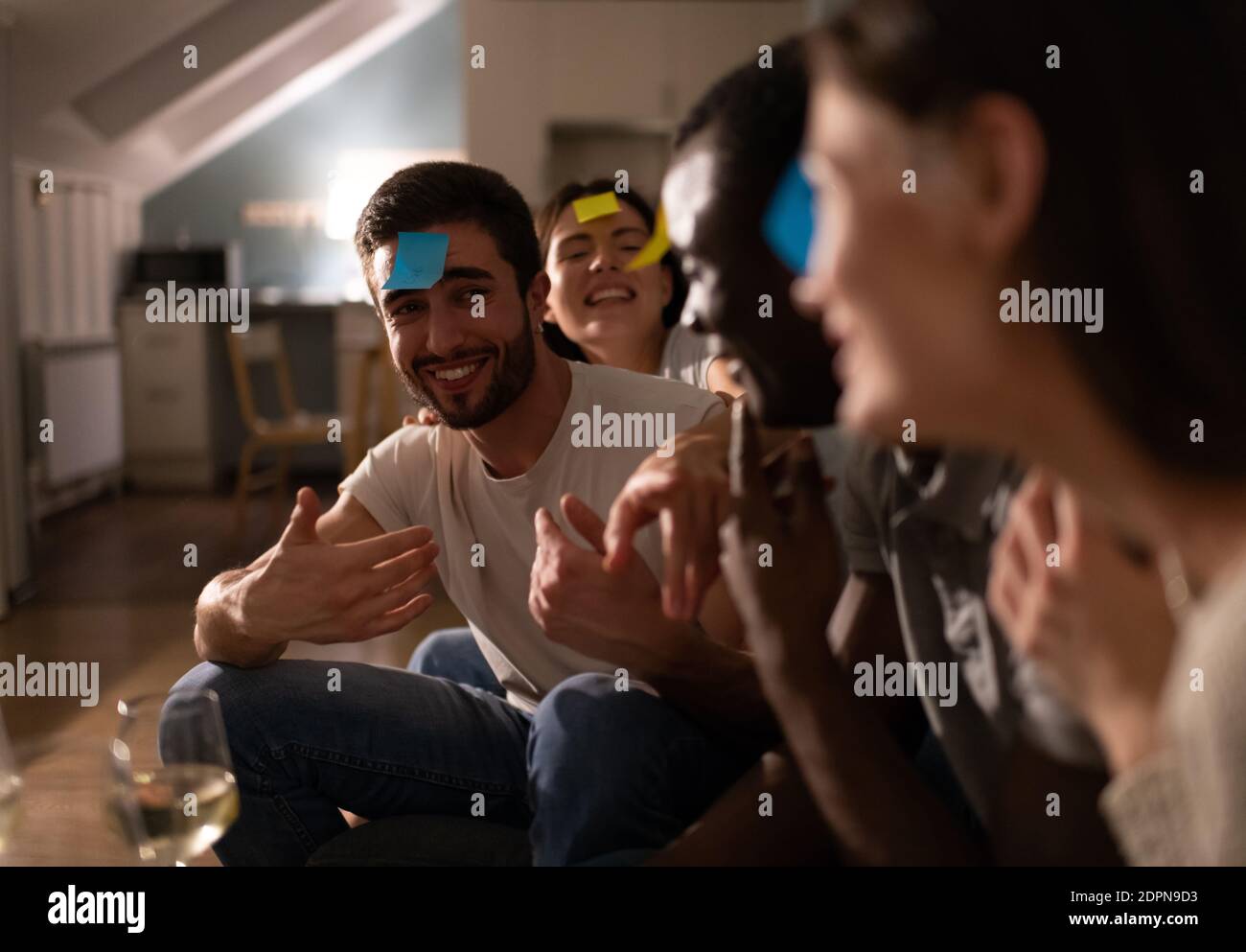 Bewildered young male with sticky note on forehead gesturing and trying to guess who is he while playing funny game with diverse friends during party Stock Photo