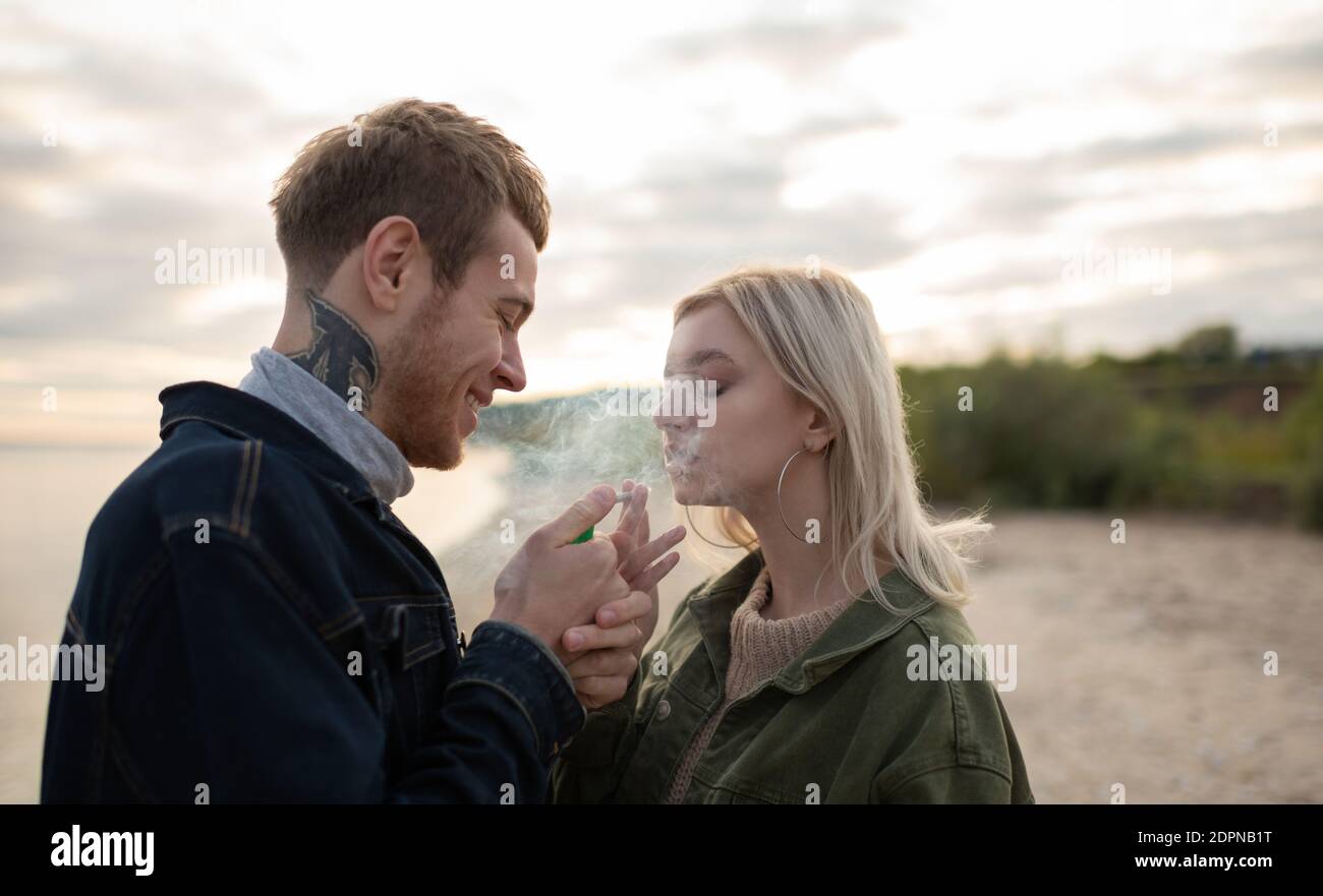 Side view of young man and woman smiling and exhaling smoke while relaxing on lake coast with cannabis cigarette Stock Photo
