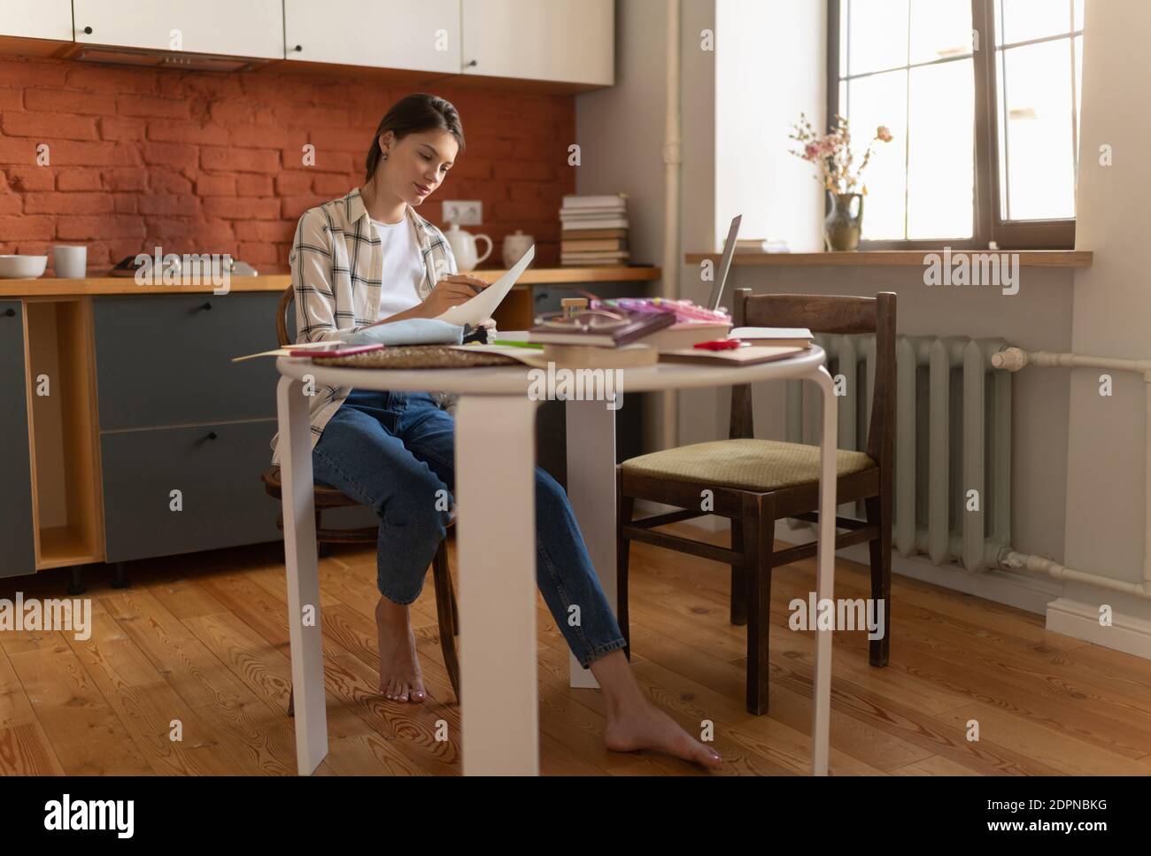 Female student in home wear preparing for exam and marking text with highlighter at table in kitchen Stock Photo