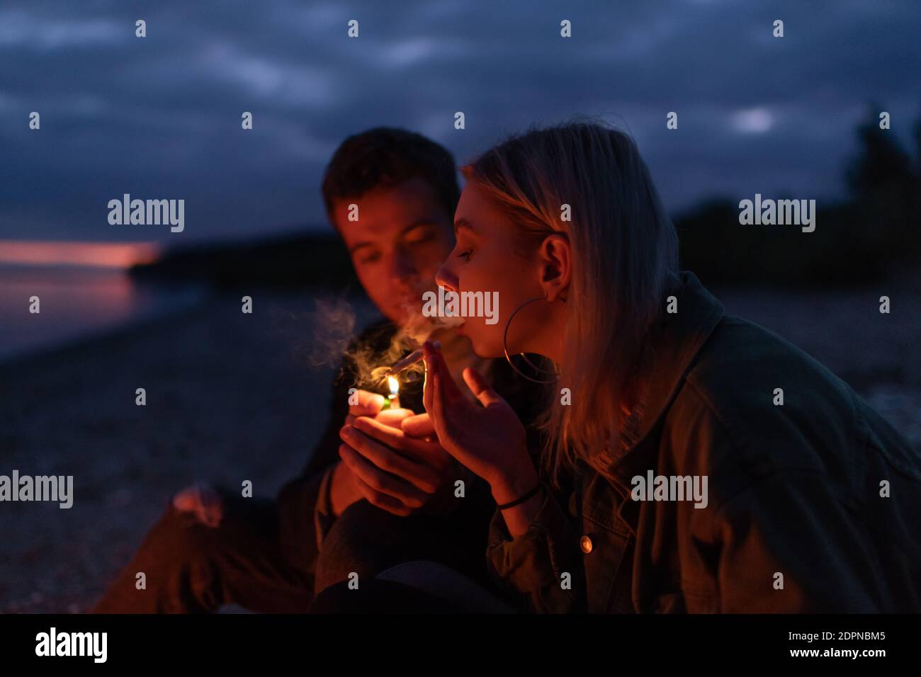 Side view of young woman exhaling smoke while boyfriend lighting weed cigarette at night on lake coast Stock Photo