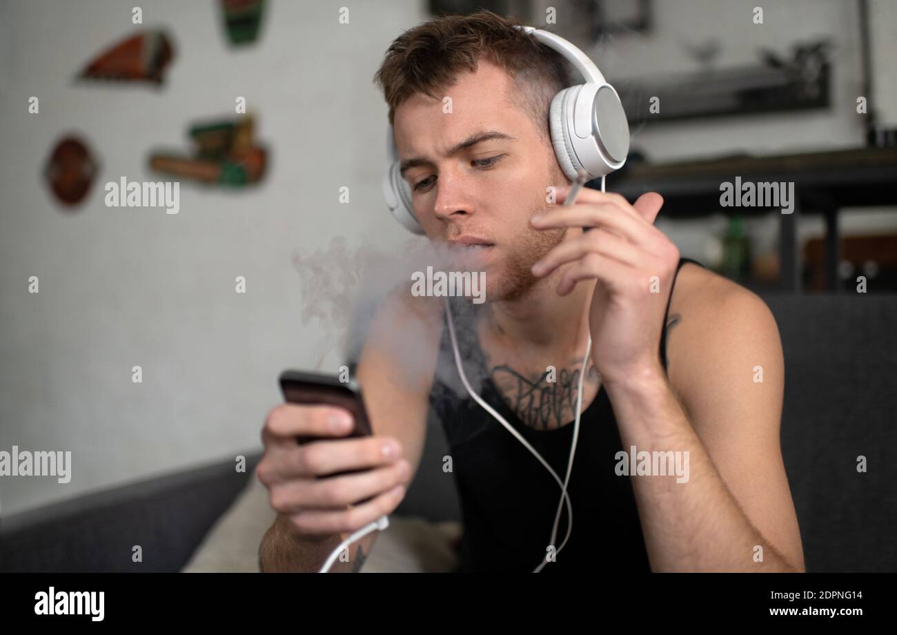 Thoughtful man with cigarette in headphones listening to music surfing  mobile phone and smoking sitting in room Stock Photo - Alamy