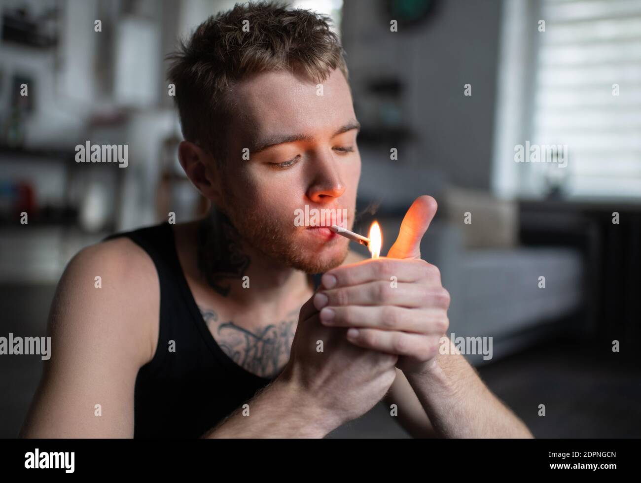 Young man inhaling smoke and igniting marijuana cigarette while sitting on blurred background of apartment Stock Photo