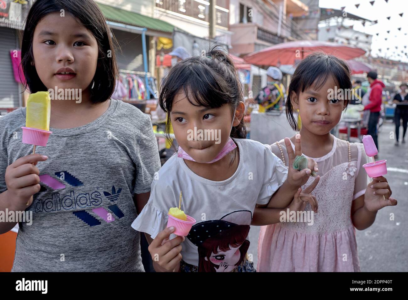 Children eating ice lollies outdoor. 3 young friends with one girl holding a fist full of money and anxious to getaway for more. Stock Photo
