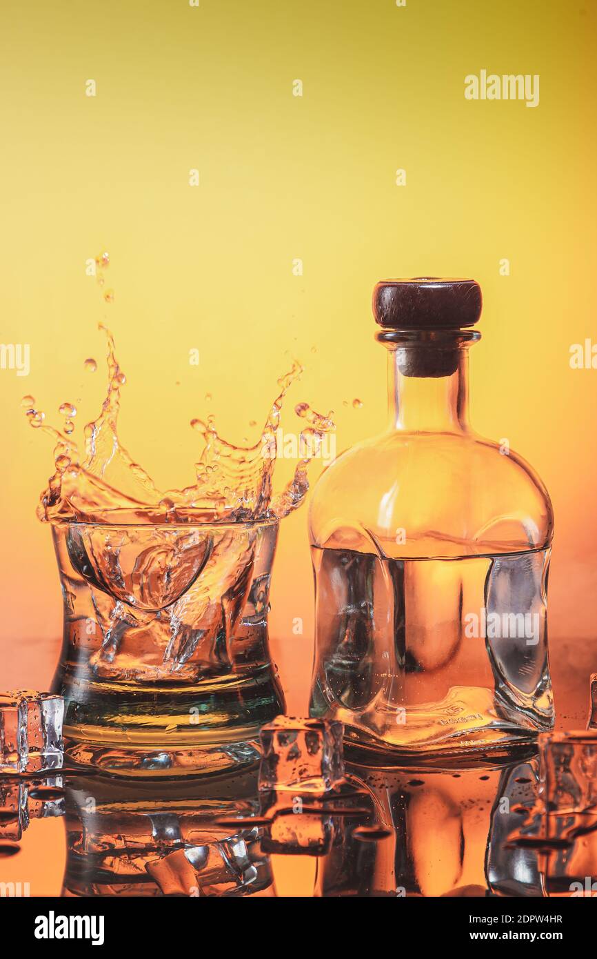 Ice falling on a glass of whisky making splash and a bottle of liqueur on orange base with ices. Whiskey and alcoholic beverages concept. Stock Photo