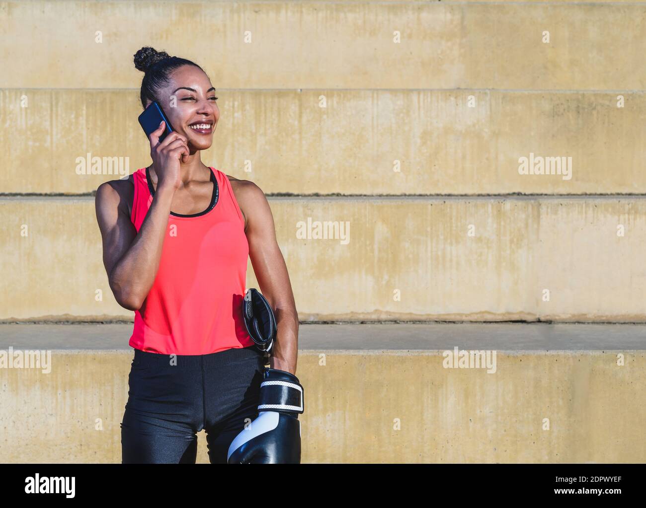 afro american attractive young woman dressing sports clothes and boxing gloves standing in a park and talking by phone while smiling. Copy space on th Stock Photo
