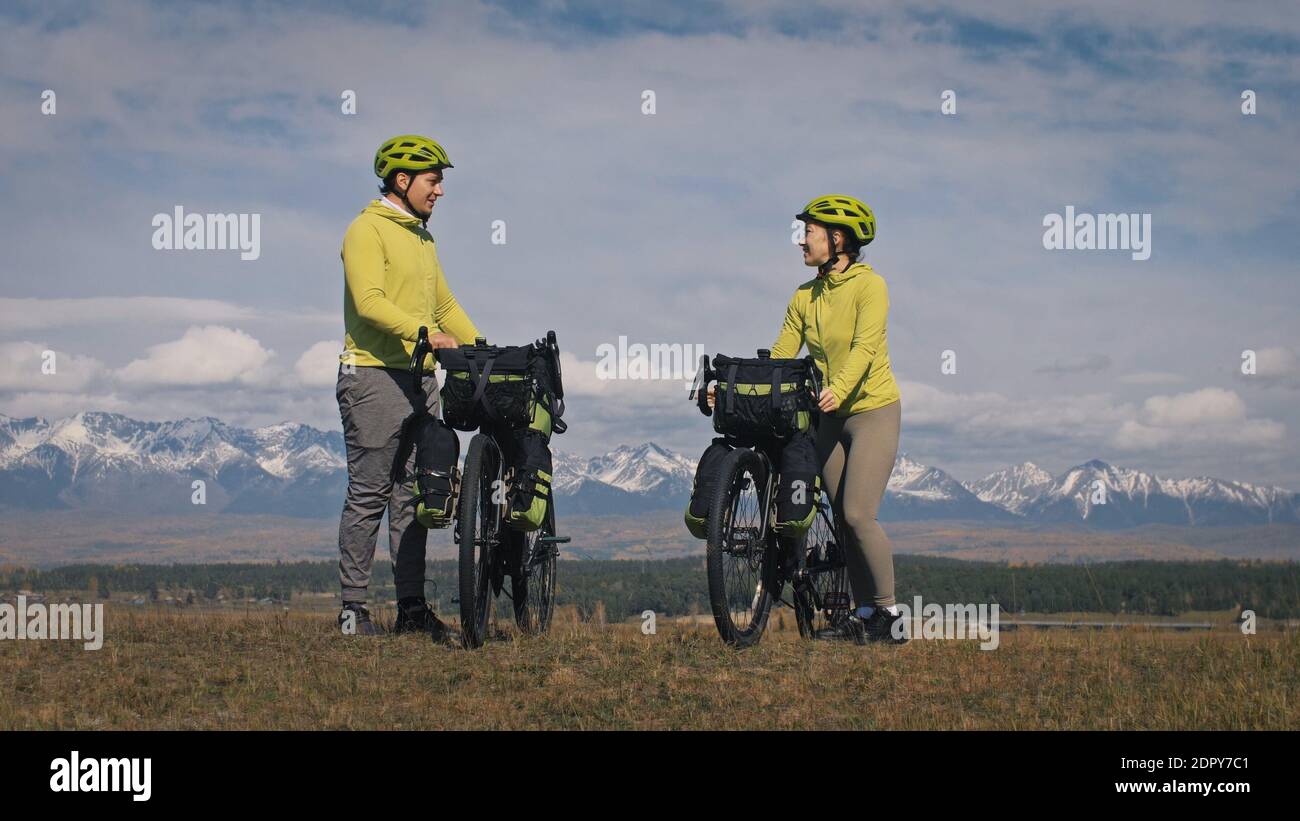 The man and woman travel on mixed terrain cycle touring with bikepacking. The two people journey with bicycle bags. Mountain snow capped. Stock Photo