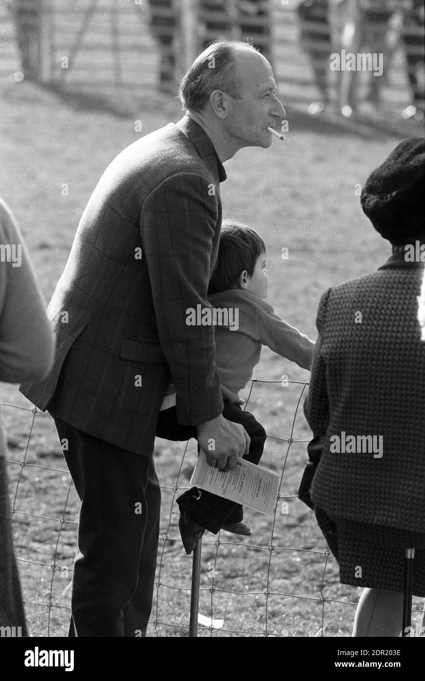 UK, England, Devonshire, Buckfastleigh, 1972. Point-to-Point races were held at  Dean Court on the Dean Marshes, close to the A38 between Plymouth and Exeter. A spectator with a boy child sitting on a shooting stick. He is smoking a cigarette and holding a racecard or racebook, a program of events. Stock Photo