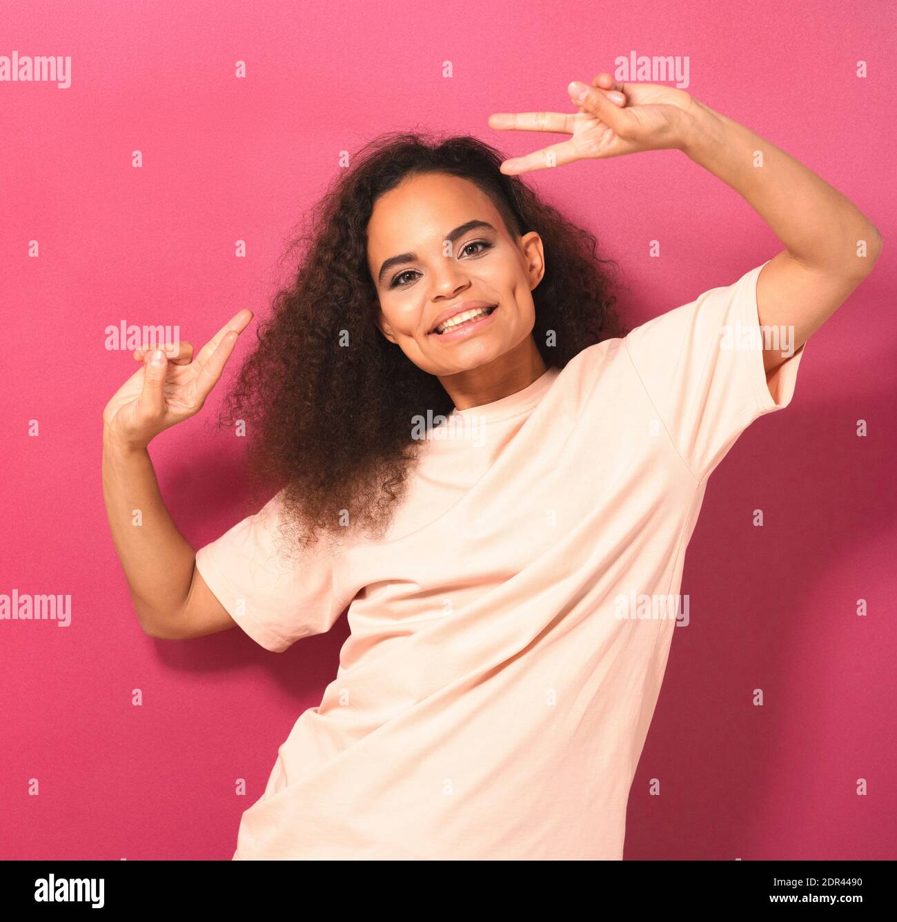 Young African American girl with Afro hair dancing gesturing peace with hands lifted up looking positively at camera wearing peachy t-shirt isolated Stock Photo