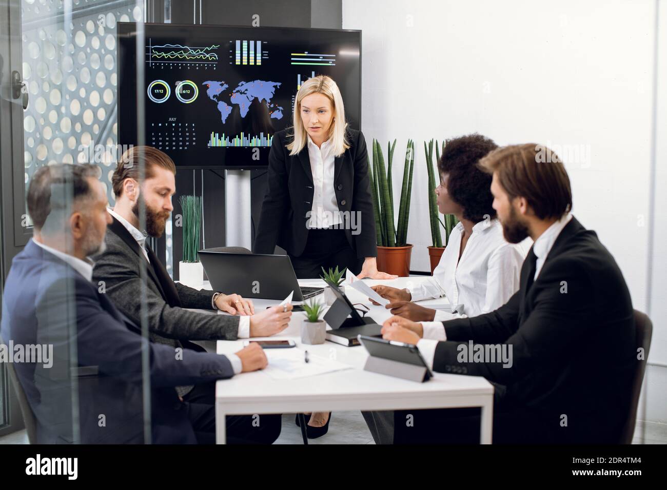 Likable concentrated blond female boss leader at a meeting together with her diverse multiracial colleagues, in modern boardroom with big TV plasma Stock Photo