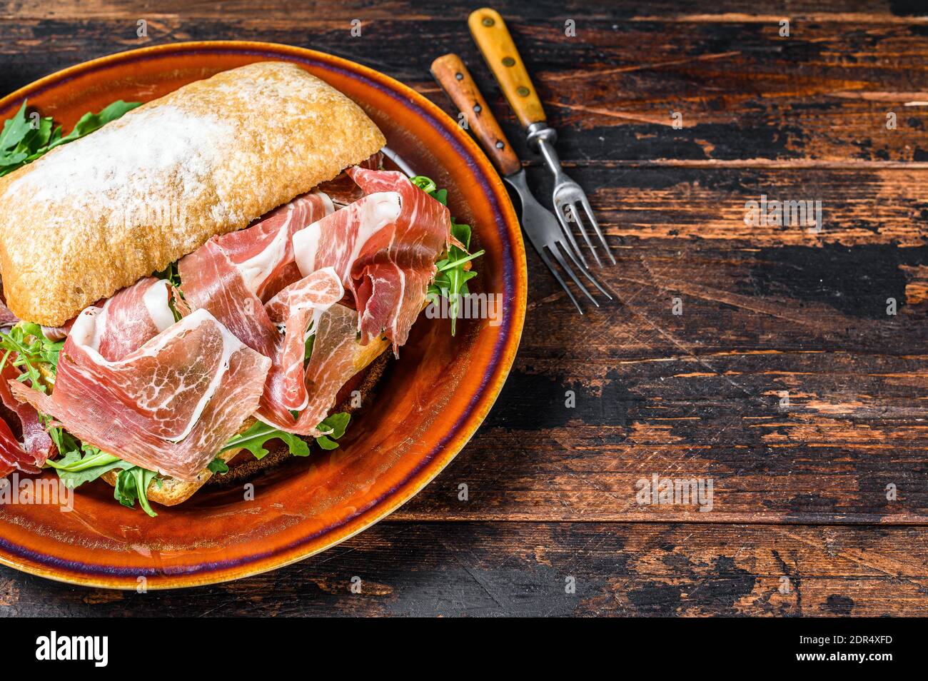 Spanish bocadillo de jamon, serrano ham sandwich on ciabatta bread with arugula. Dark wooden background. Top view. Copy space. Stock Photo
