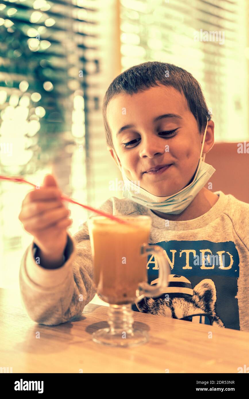 A child wearing a mask in a cafe during a pandemic. A boy in a cafe drinks sea buckthorn tea in a cafe during a pandemic. toned. vertical photo Stock Photo