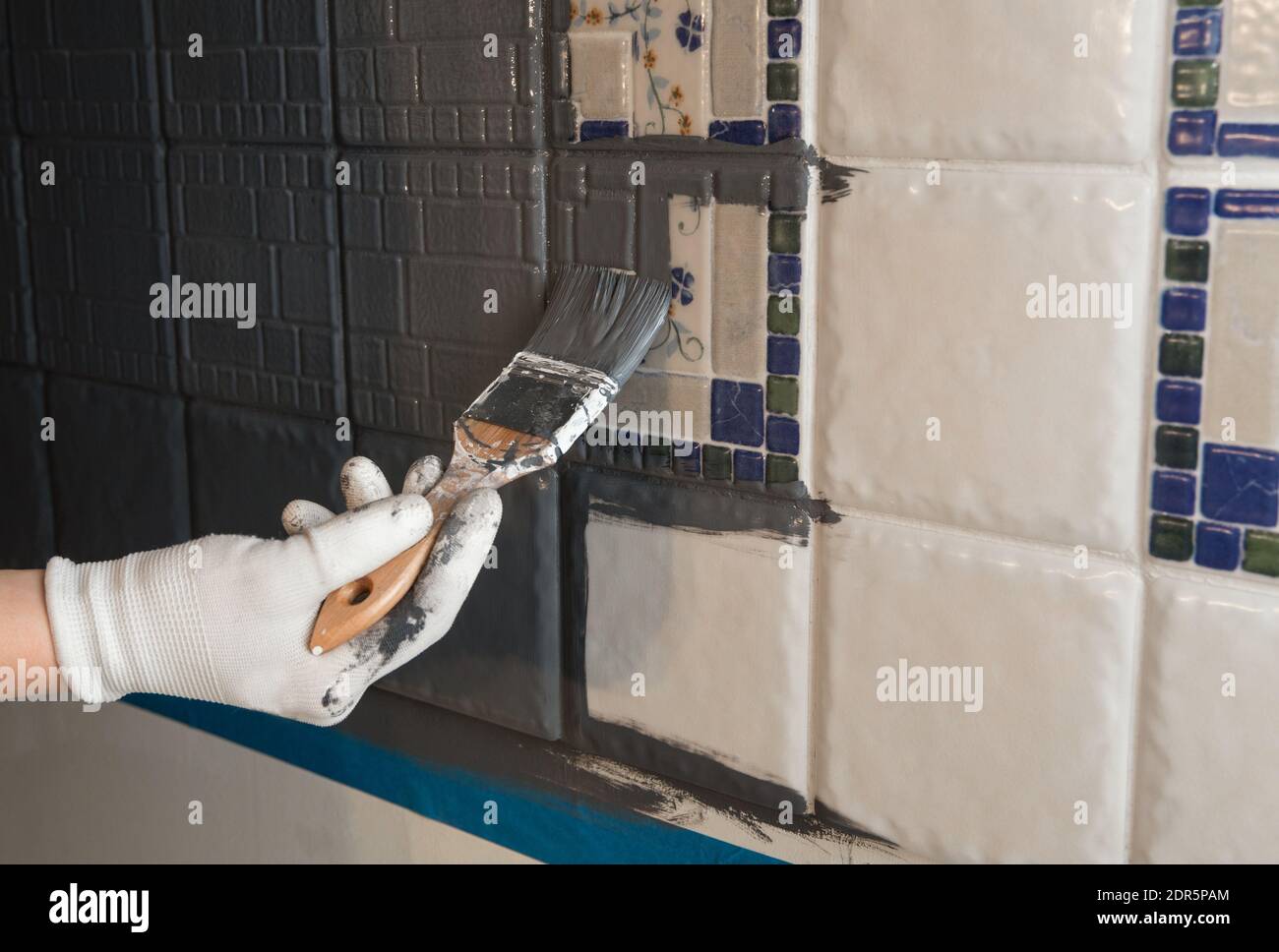 Repainting old dated kitchen ceramic tile back wall with modern gray chalk paint indoors at home. Giving old kitchen new look concept. Hand holding a Stock Photo