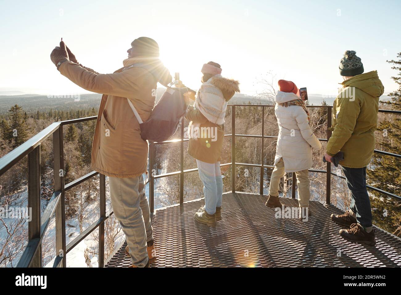 Group of young friends in winterwear taking photo of majestic scenery of mountains and forest covered with snow against sun shining on blue sky Stock Photo