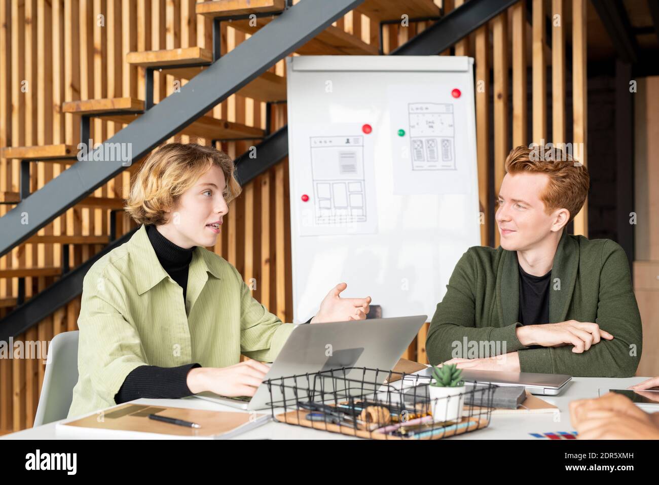 Young blond female mobile app developer looking at one of her colleagues during brainstorm and discussion of creative ideas at meeting Stock Photo