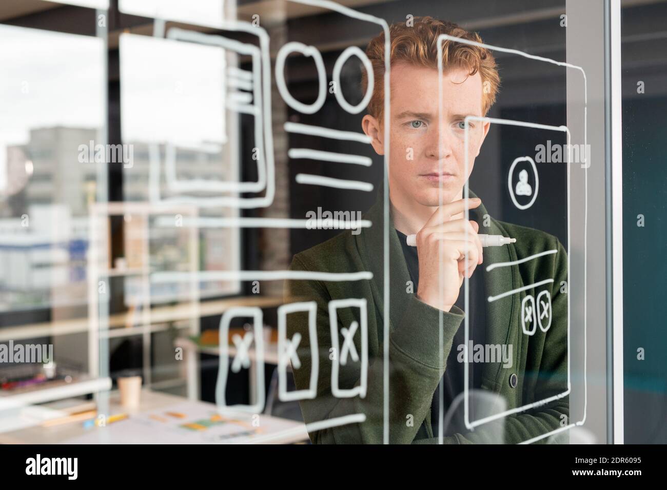 Young pensive male software developer keeping hand by chin while standing in front of noticeboard and looking at sketches of mobile app Stock Photo