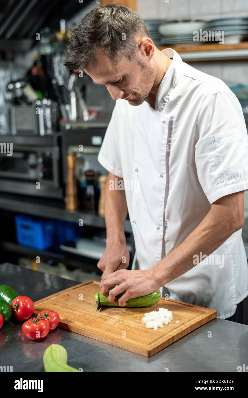 Young male chef in uniform bending over table while cutting fresh green zucchini on wooden board surrounded by tomatoes and avocado Stock Photo