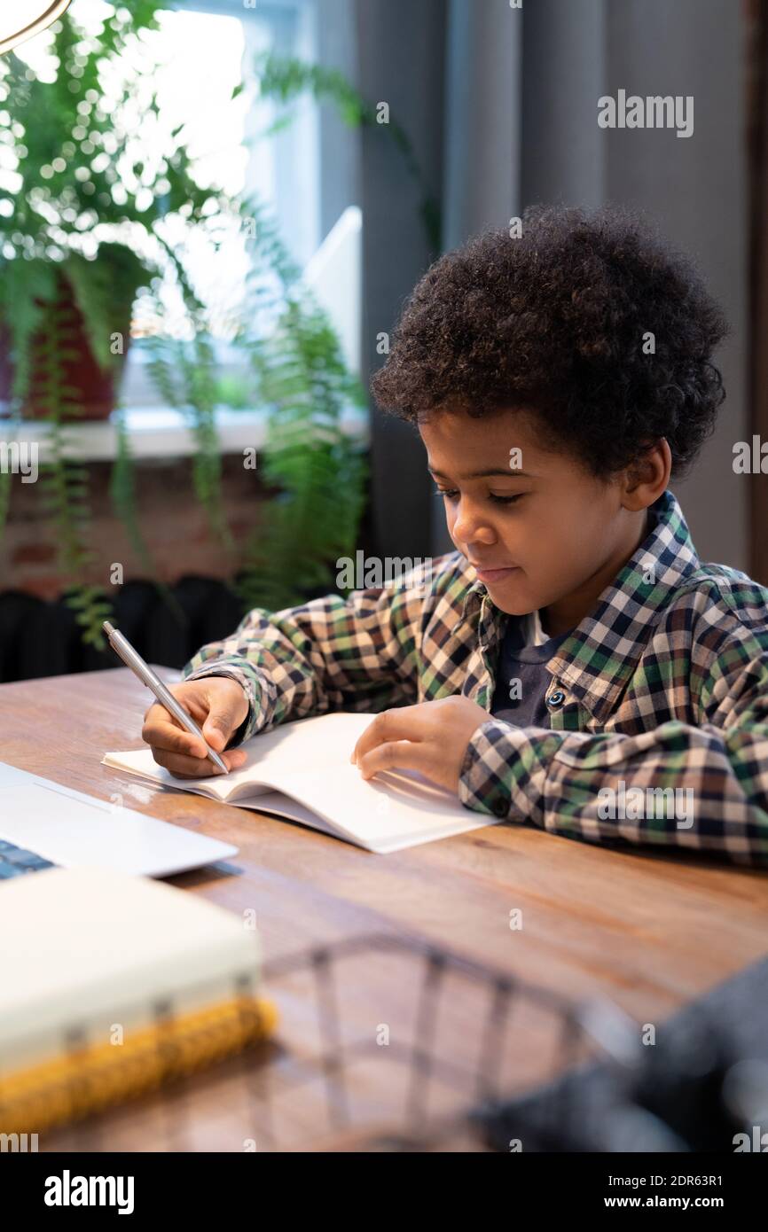 Cute little African schoolboy with pen making notes on page of copybook ...