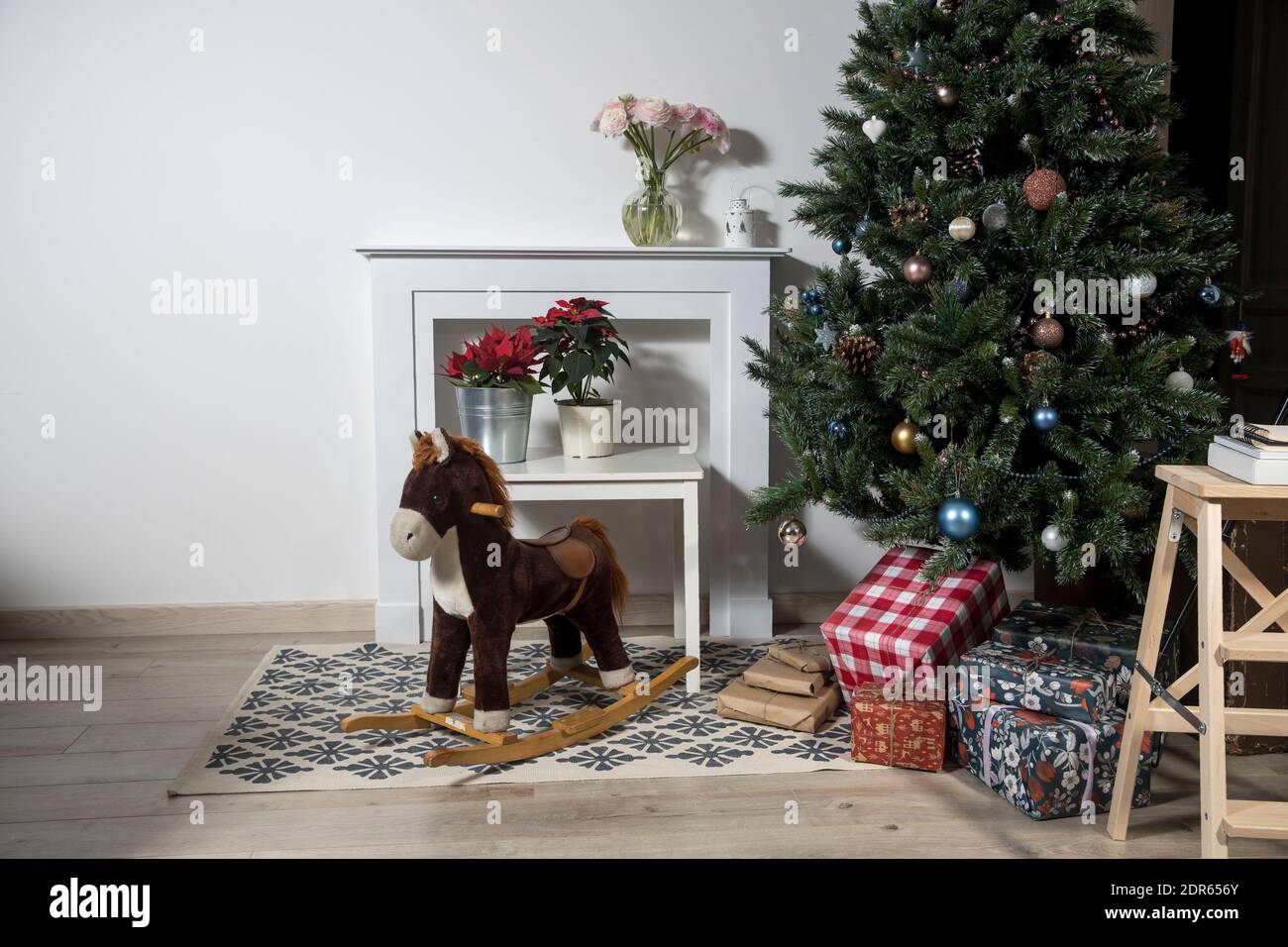 Interior with Christmas tree decorated, white fake fireplace with flowers and a rocking horse. Paper garland on the wall. The boses with gifts. Stock Photo