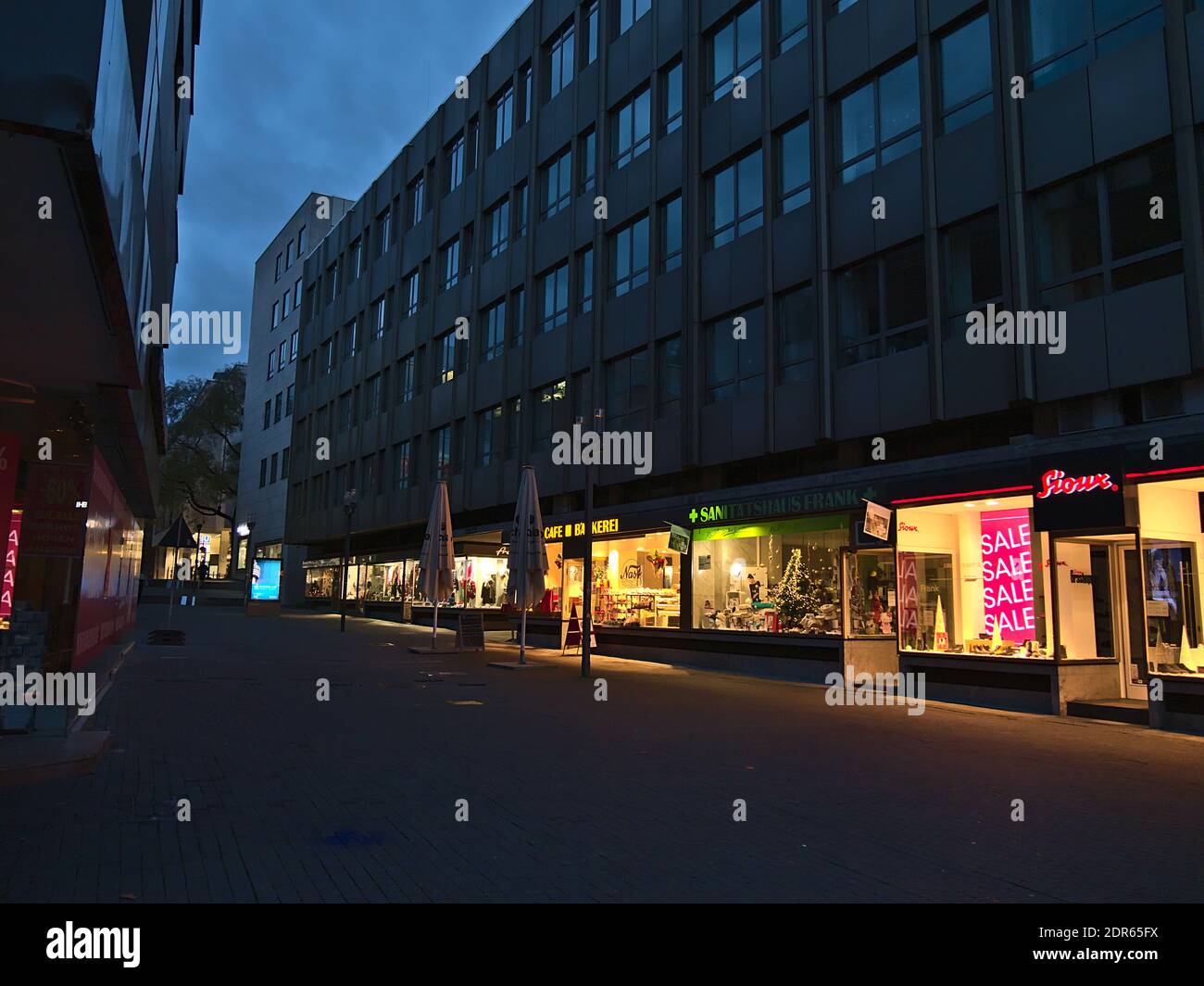 Deserted shopping street in city center with row of closed stores during Covid-19 lockdown and illuminated shop windows in evening in Christmas season. Stock Photo