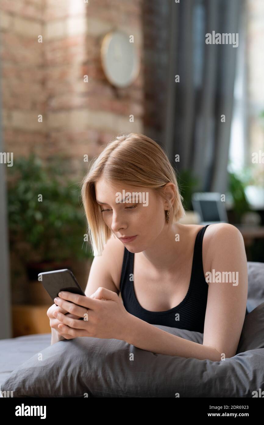 Young restful blond female with smartphone texting or scrolling through online news while relaxing on bed in bedroom on Sunday morning Stock Photo