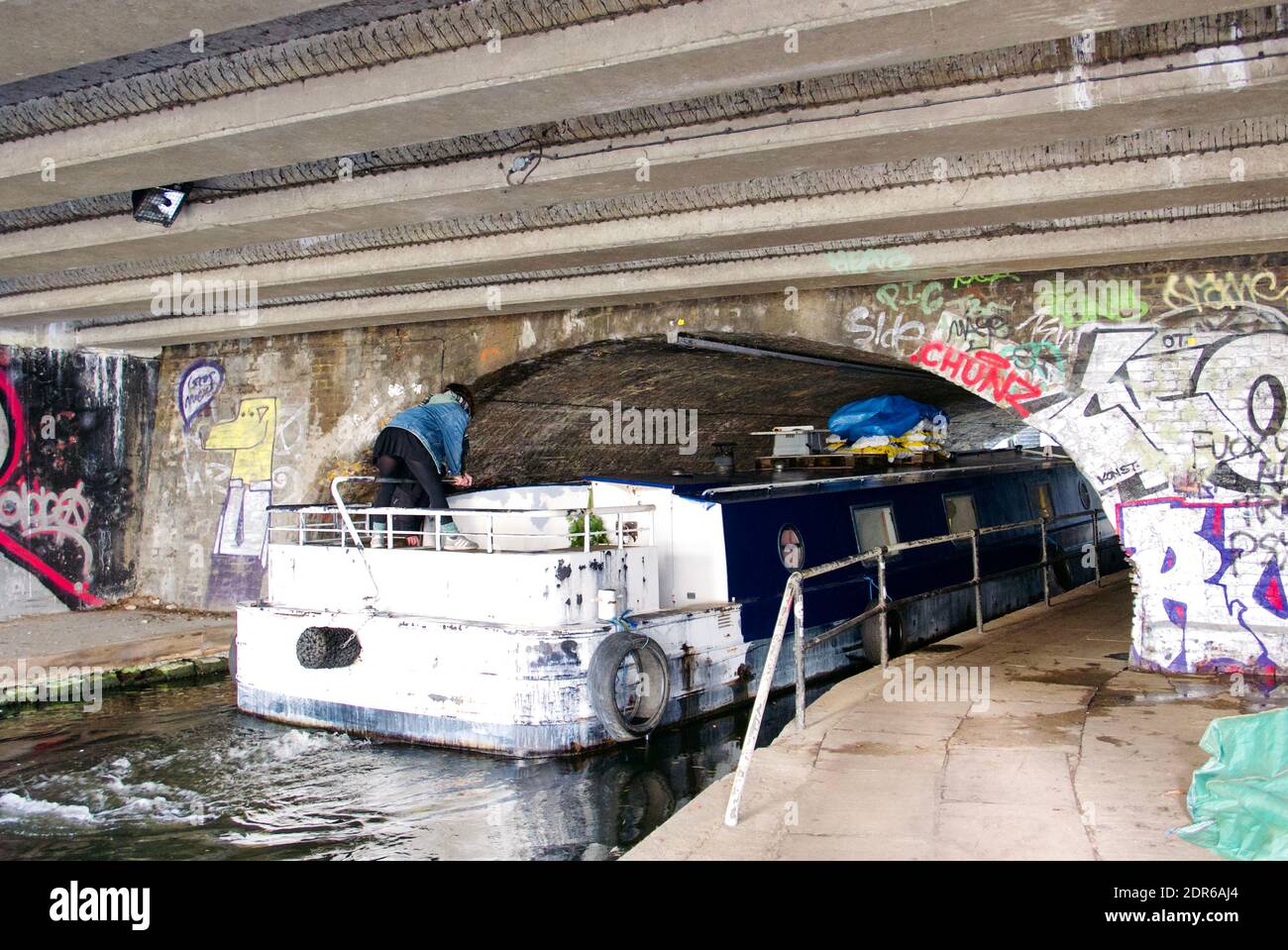 Young woman wearing denim jacket and skirt steers her canal boat through a tunnel approaching Kentish Town canal lock on Regents Canal, London. Stock Photo