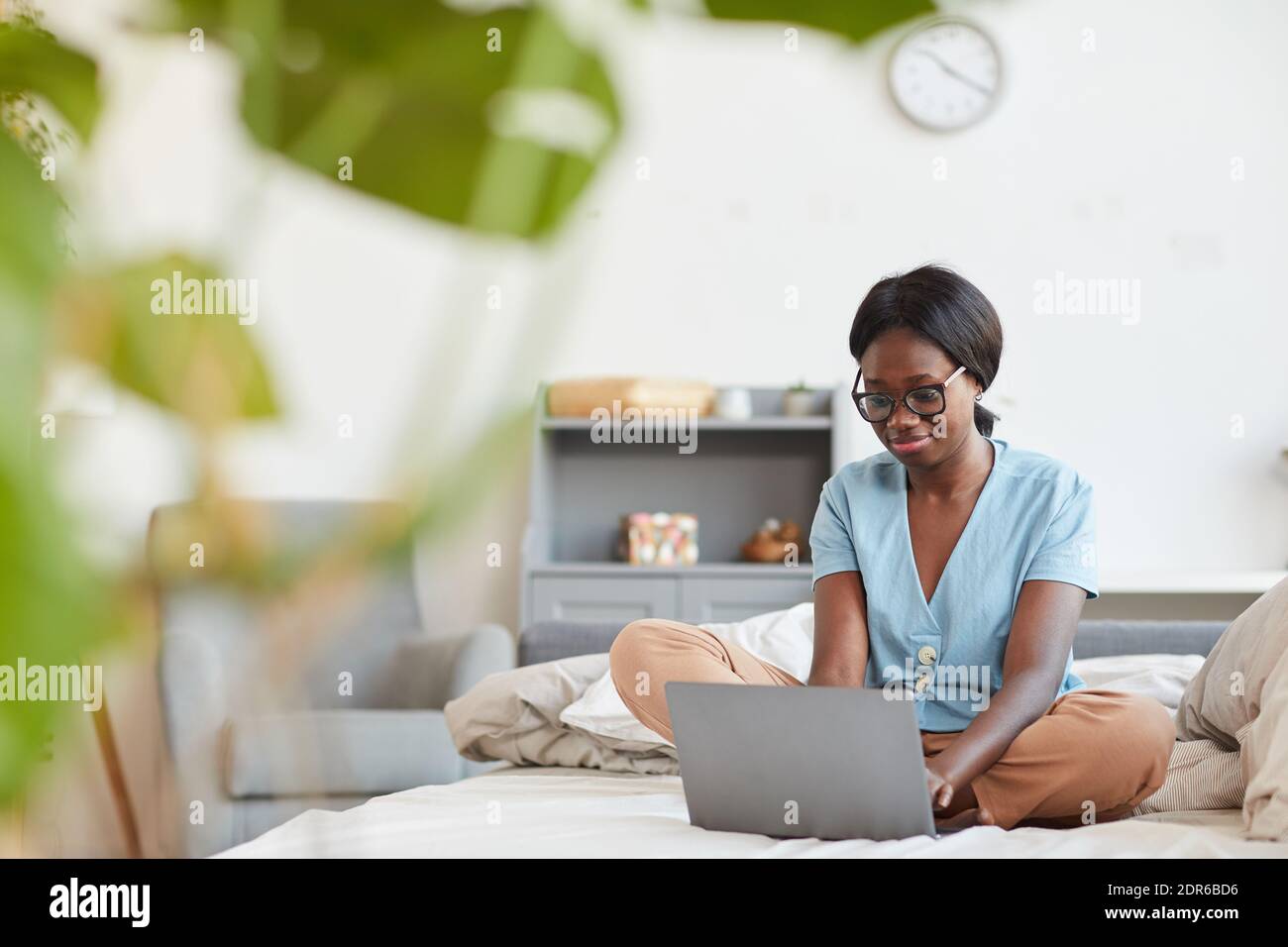Full length portrait of young African-American woman using laptop while sitting on bed at home with plant leaves framing, copy space Stock Photo