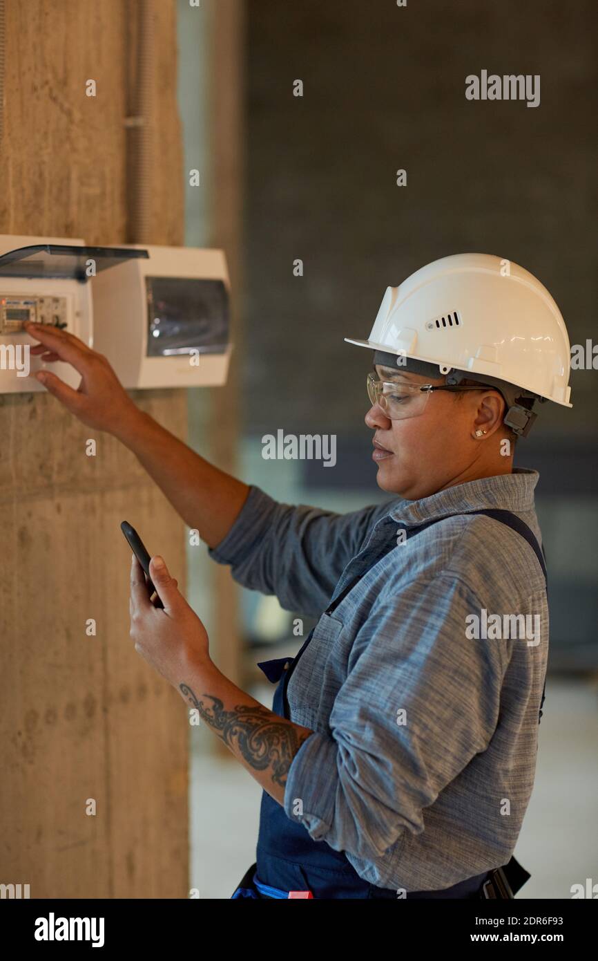 Vertical side view portrait of female worker setting up electricity and using smartphone while working on construction site Stock Photo