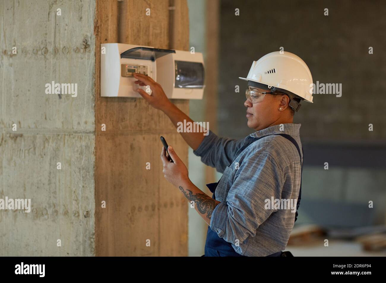 Side view portrait of female worker setting up electricity and using smartphone while working on construction site, copy space Stock Photo