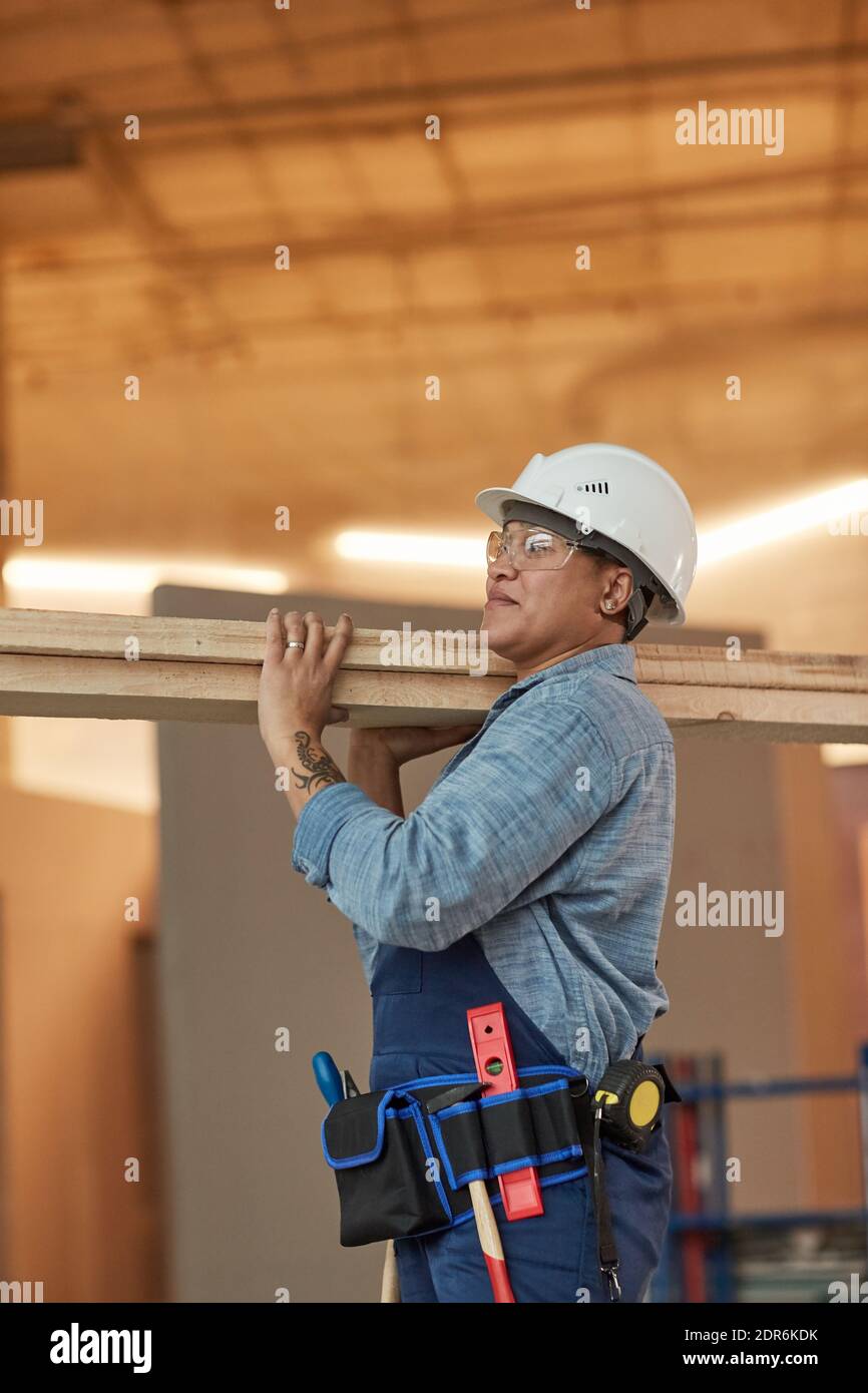 Vertical side view portrait of modern female worker carrying wood boards while working on construction site Stock Photo