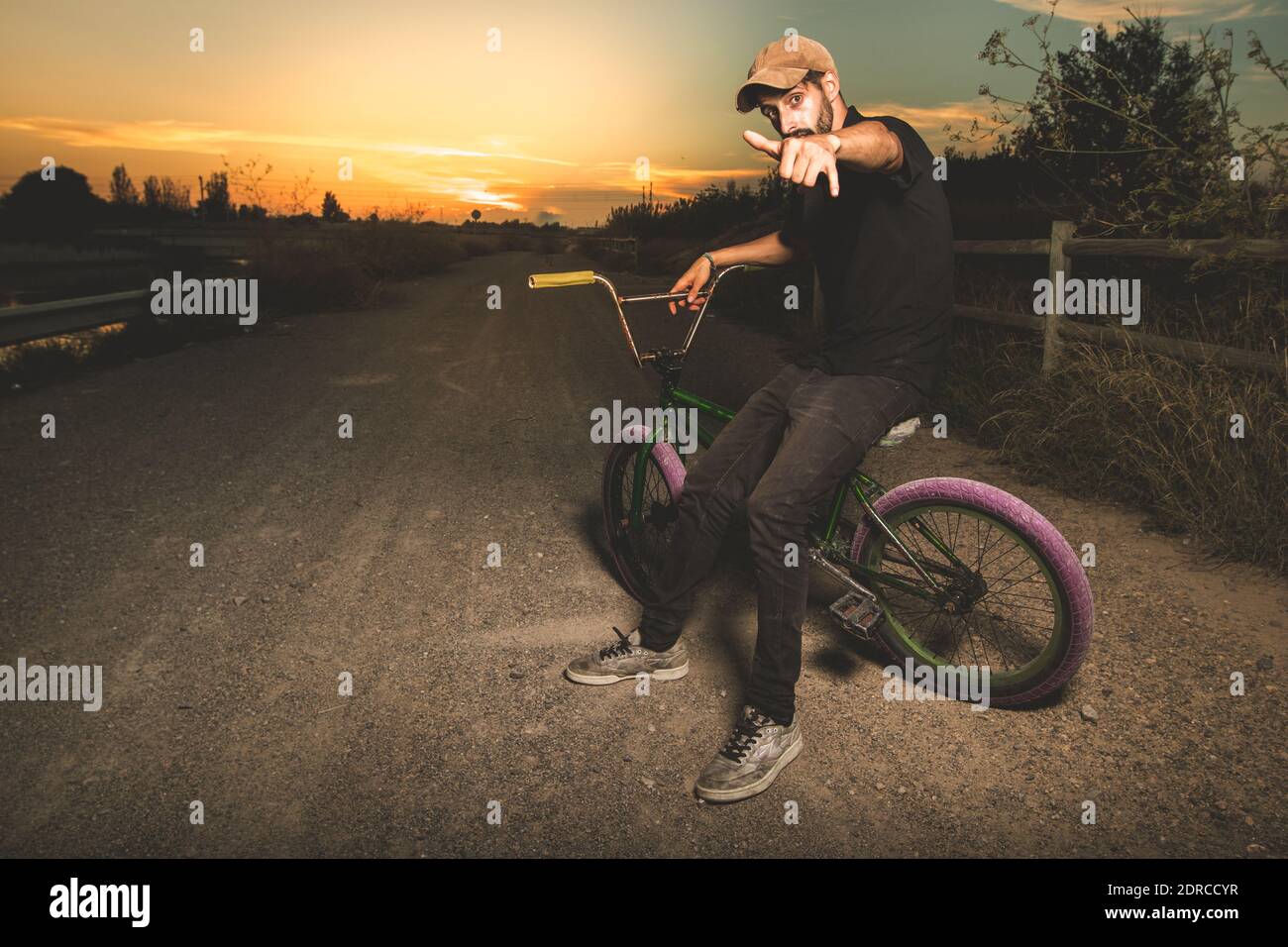 Portrait Of Man Gesturing Shaka Sign While Sitting On Bicycle Against Sky During Sunset Stock Photo