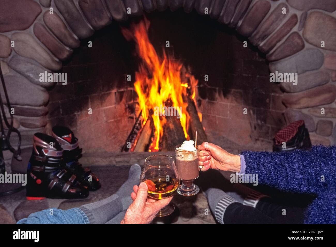Two relaxed downhill skiers sit by a fireplace with their boots off and in stocking feet to toast each other with après-ski drinks and warm up in front of a blazing wood fire in a ski lodge at Big Sky Resort in Montana, USA. Brandy and hot chocolate are among America's most popular after-ski drinks, along with mulled wine, Irish coffee, hot buttered rum, and spiced apple cider. Big Sky is the largest of all ski resorts in that  Northwestern state and boasts 31 ski lifts that give easy access to Rocky Mountain slopes with more than 300 ski runs for beginning, intermediate and expert skiers. Stock Photo