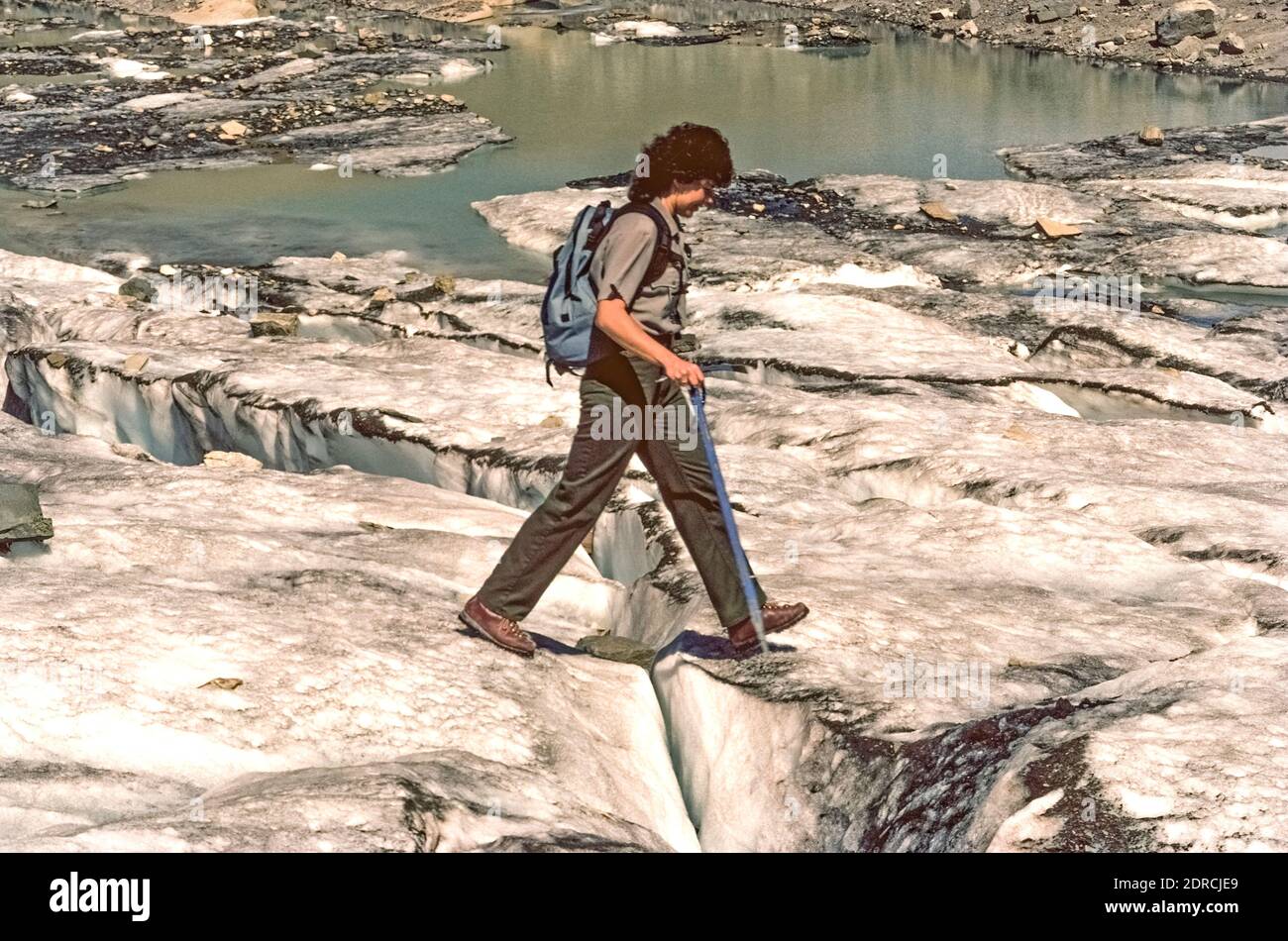 Using an ice axe for safety and support, a hiking female ranger with the United States National Park Service (NPS) steps across a fissure in the icy remnant of the receding Grinnell Glacier, a longtime attraction in Glacier National Park in northwestern Montana, USA. The glacier was named for George Bird Grinnell, an early conservationist who discovered the ice field in 1885 and helped the pristine region to be established as America's 10th national park in 1910. Unfortunately the numerous glaciers for which the park is named have suffered from climate change over the years. Stock Photo