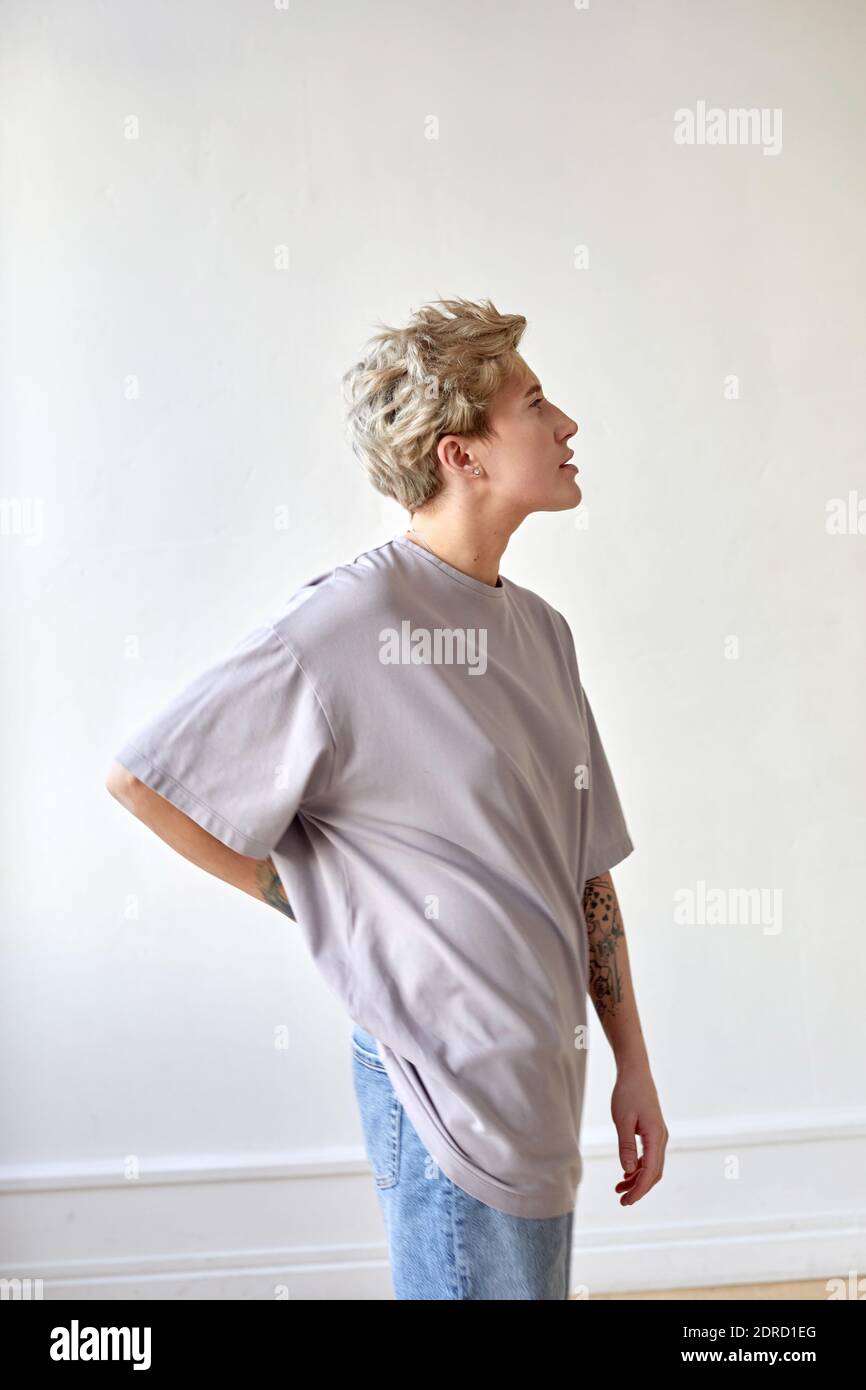 Side view of androgynous female in baggy t shirt and jeans holding hand on waist and looking away against white wall at home Stock Photo